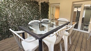 Un patio al aire libre con una mesa de vidrio rodeada de sillas blancas, una terraza de madera y una pared cubierta de follaje verde.