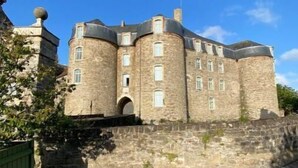Un castillo histórico de piedra con múltiples torres y ventanas arqueadas, rodeado por un muro de piedra y un camino adoquinado.