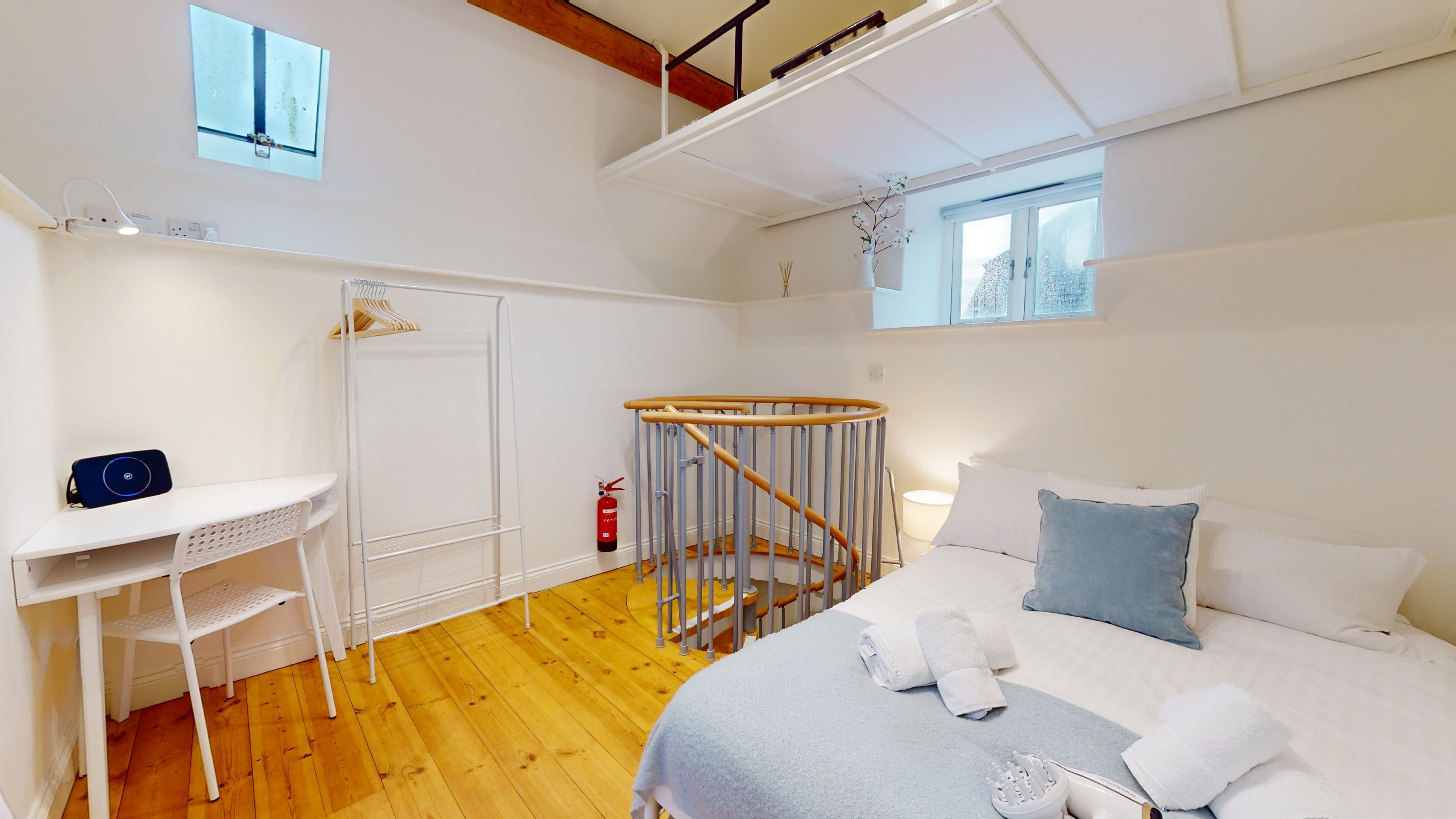 A bedroom with a bed, a desk, a chair, a wooden floor, a window, and a ceiling light.