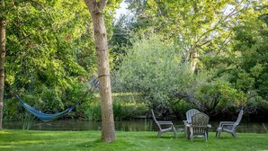 A hammock between trees, two Adirondack chairs, and a grassy area near a body of water.