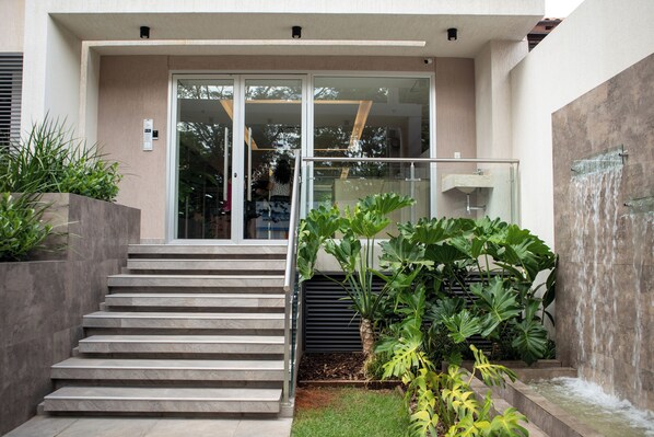 A modern building entrance with a glass door, a staircase, and a small garden area with large plants.