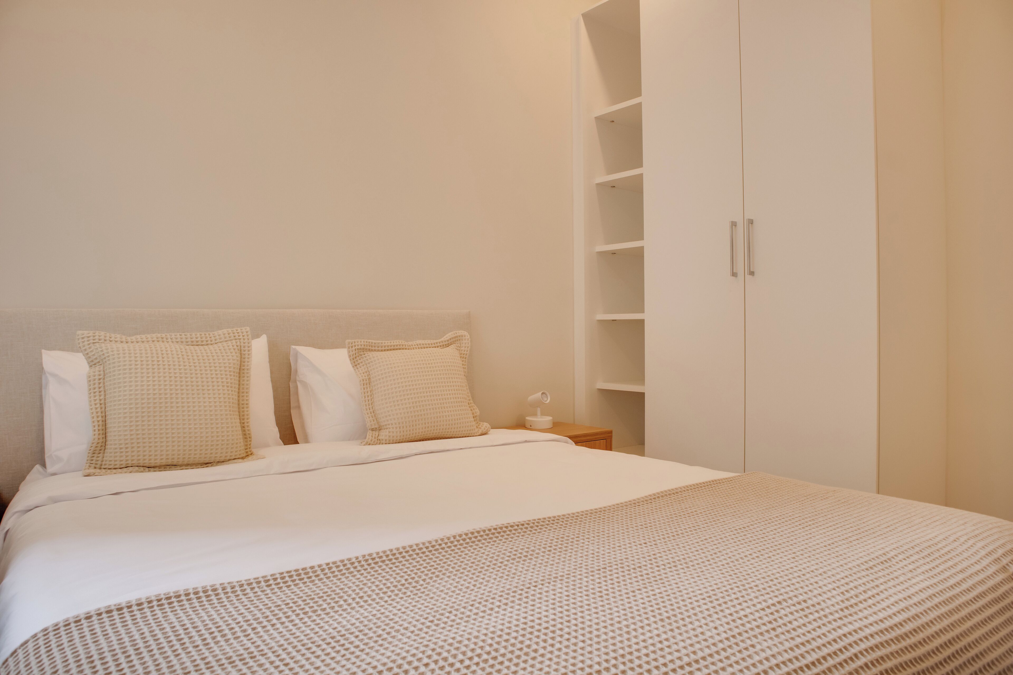 Ein ordentlich bezogenes Bett mit einem gemusterten Überwurf und zwei Kissen, ein Nachttisch mit Lampe sowie ein weißer Kleiderschrank mit mehreren Regalböden.
