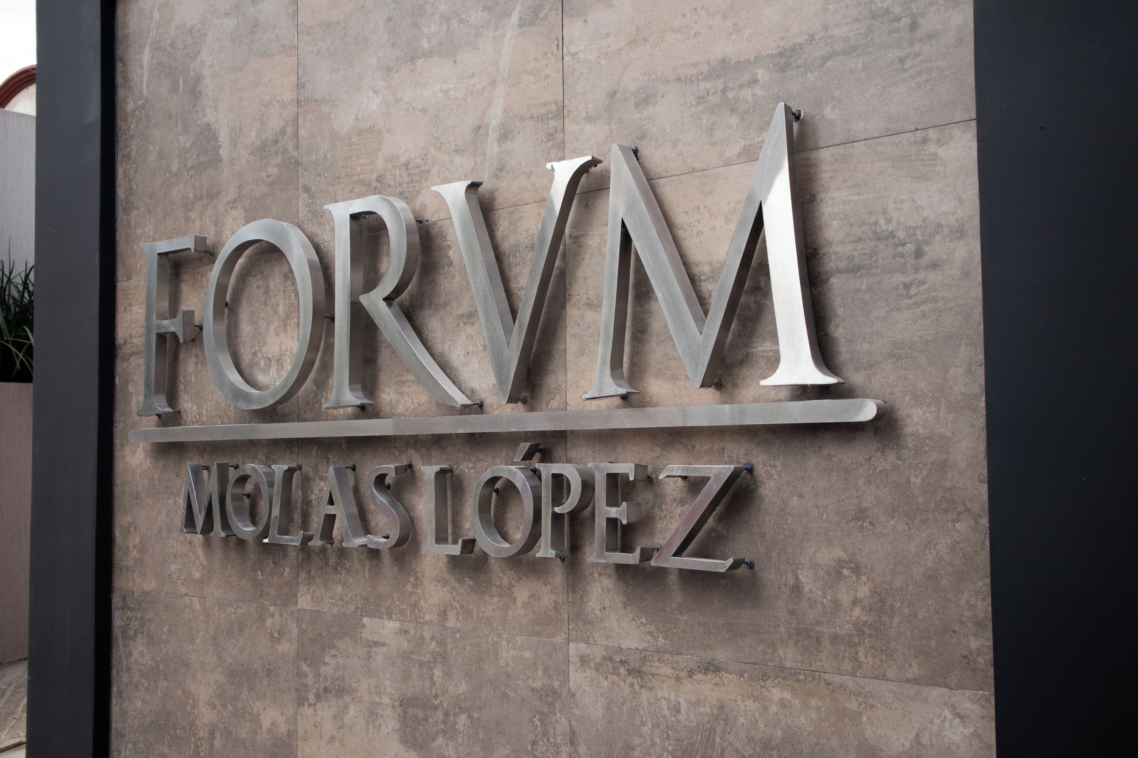 A metallic sign with the text "FORVM MELAS LOPEZ" mounted on a stone wall.