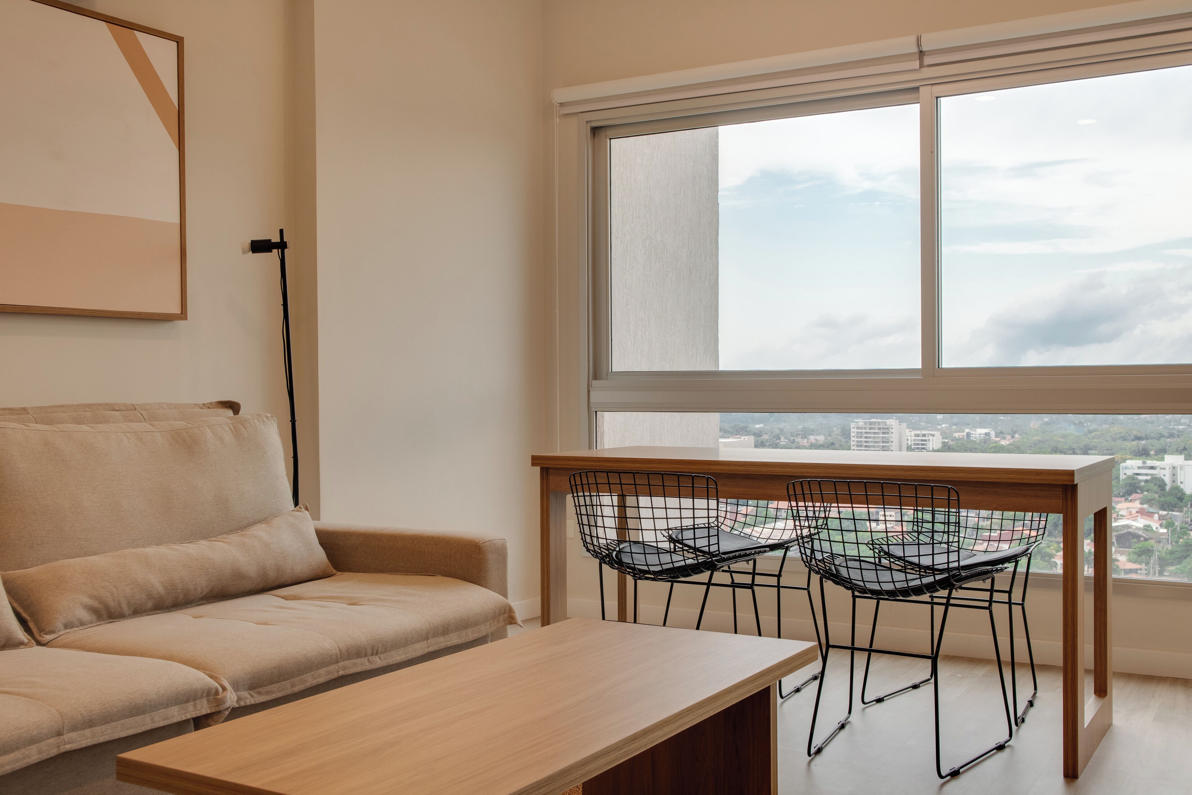 A modern living room with a beige sofa, a wooden coffee table, and a dining table with wire chairs. A large window offers a view of the cityscape.