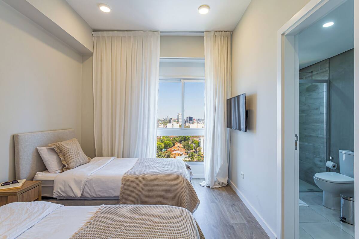 A hotel room with two beds, a TV, a window with curtains, and a view of the city.