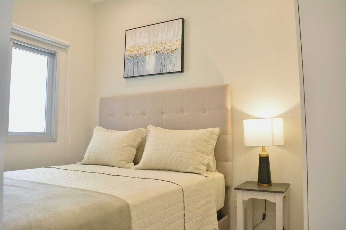 A bedroom with a bed, two pillows, a nightstand with a lamp, and a painting on the wall.
