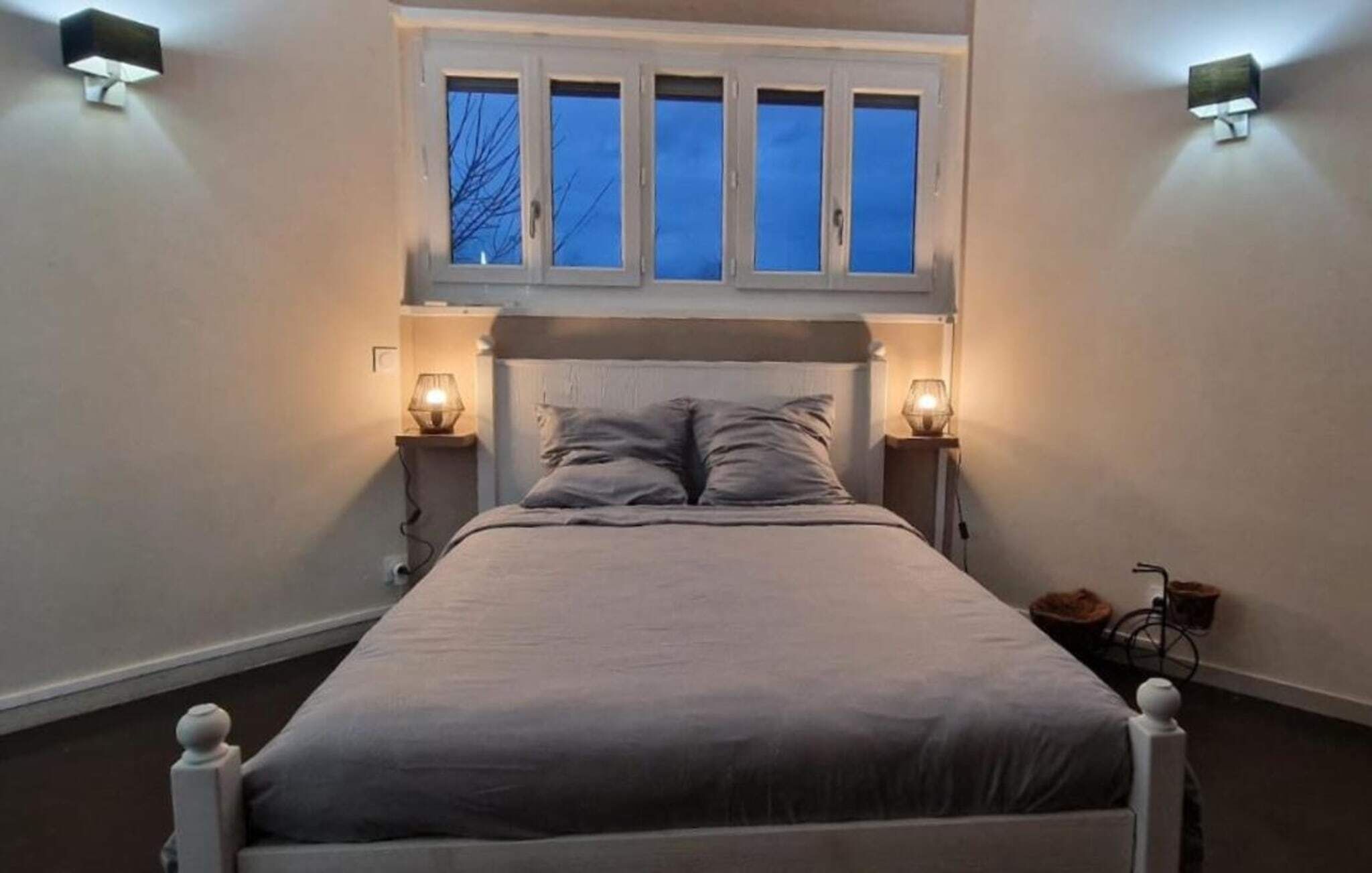 A bedroom with a bed, two bedside lamps, a window with blinds, and a wall-mounted light fixture.