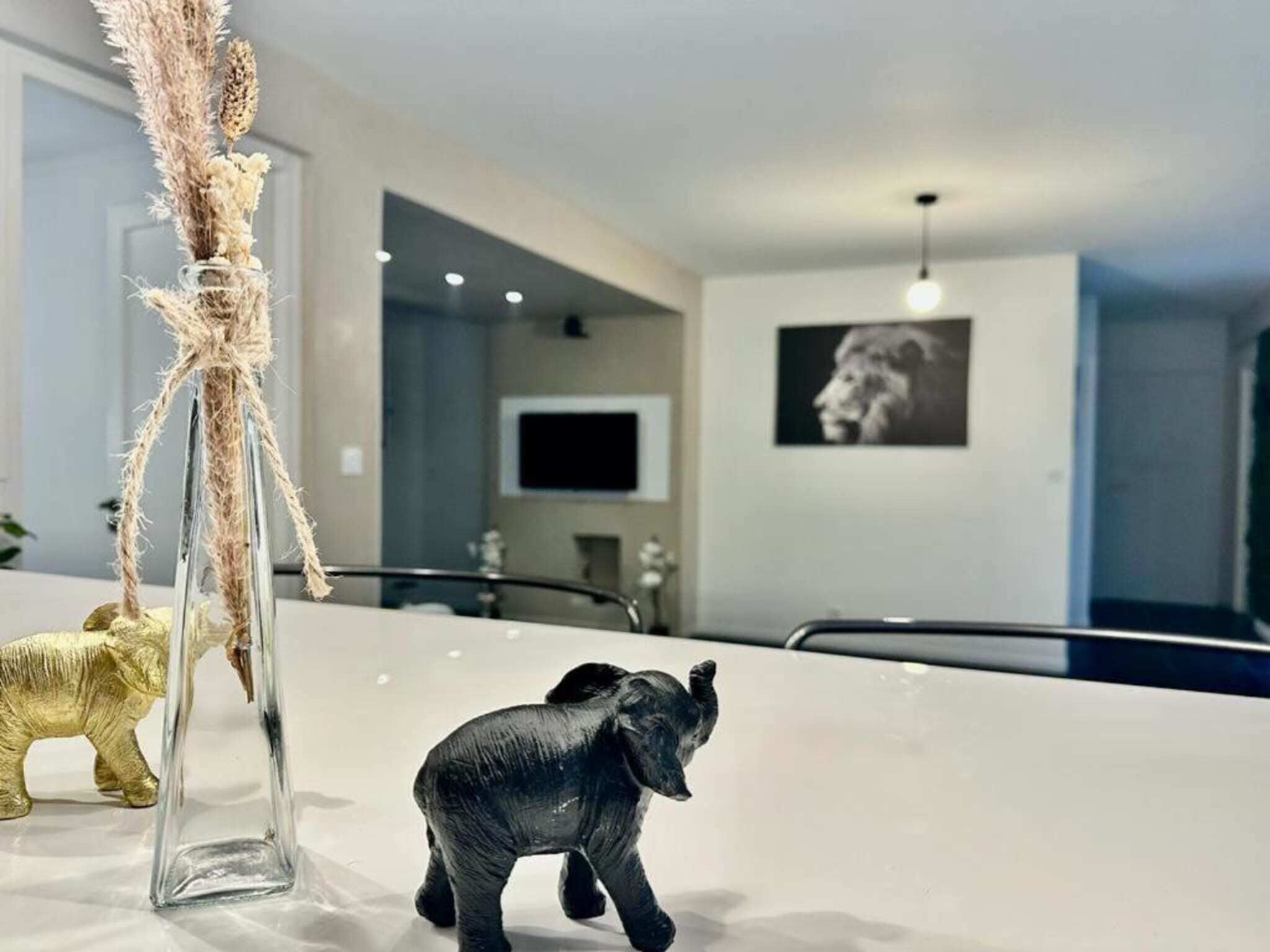 A modern interior with a clear glass vase with dried flowers, a small elephant figurine, and a golden elephant statue on a white surface.