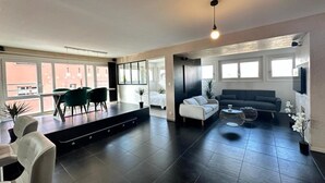 A spacious living area with a dark tiled floor, a sofa, a coffee table, and a dining area with chairs and a table. - L' Elegant (Douvaine)