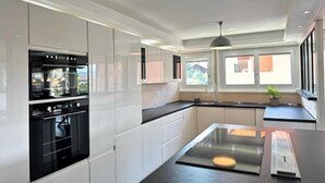 A modern kitchen with white cabinetry, a built-in oven, a central island, and a window providing natural light. - L' Elegant (Douvaine)