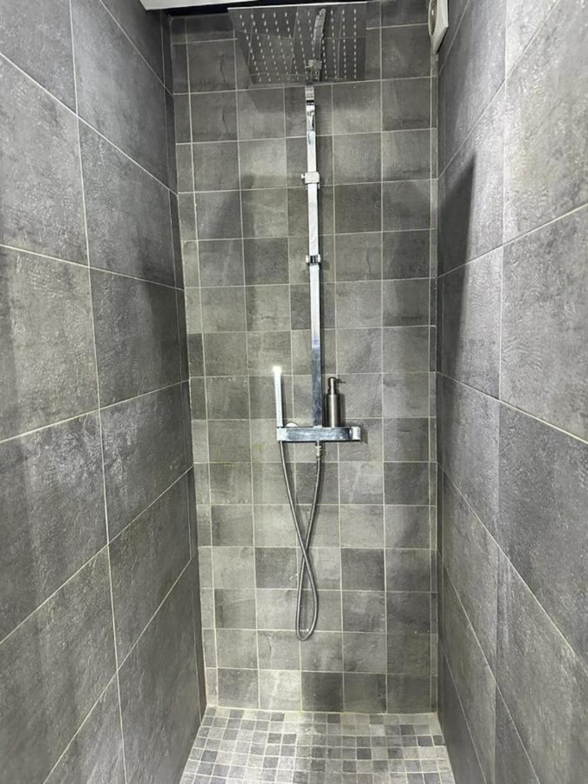 Un vestuario de ducha pequeño, con azulejos y un cabezal de ducha manual, mostrando un chorro de agua vertical.