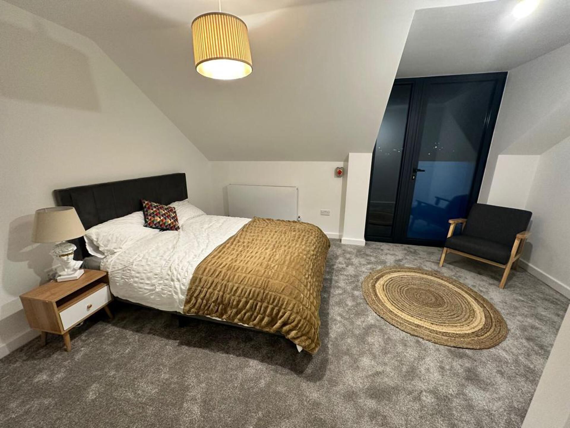 Un dormitori amb llit, taula de nit, làmpada i vista a una habitació fosc per una porta.