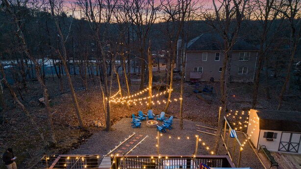 A deck with outdoor seating and string lights, surrounded by trees and a building in the background. - Walk to Beach HotTub FirePit Game room Lake View (East Stroudsburg)