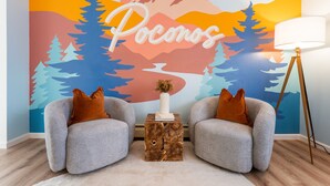 A room with two grey armchairs, a small wooden side table with a vase, and a floor lamp. A large wall mural with the word "Pocanos" and a mountain scene is visible. - Walk to Beach HotTub FirePit Game room Lake View (East Stroudsburg)