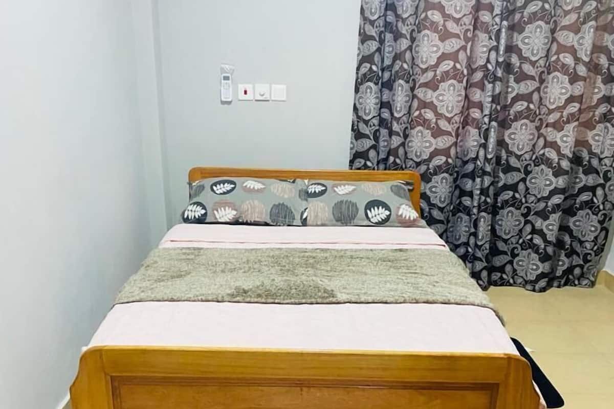 A wooden bed with a white mattress and a grey blanket, positioned against a wall with a patterned curtain.