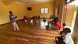 A room with wooden flooring and a group of people sitting in a circle, facing a person standing at the front. - Akpe Cultural Center and Hotel (Accra)