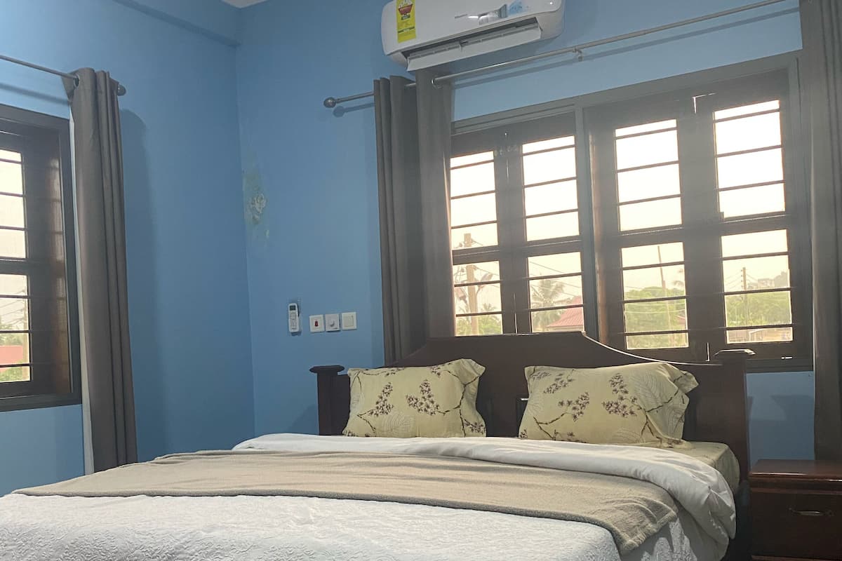 A bedroom with a bed, an air conditioner, a nightstand, and a window with curtains.