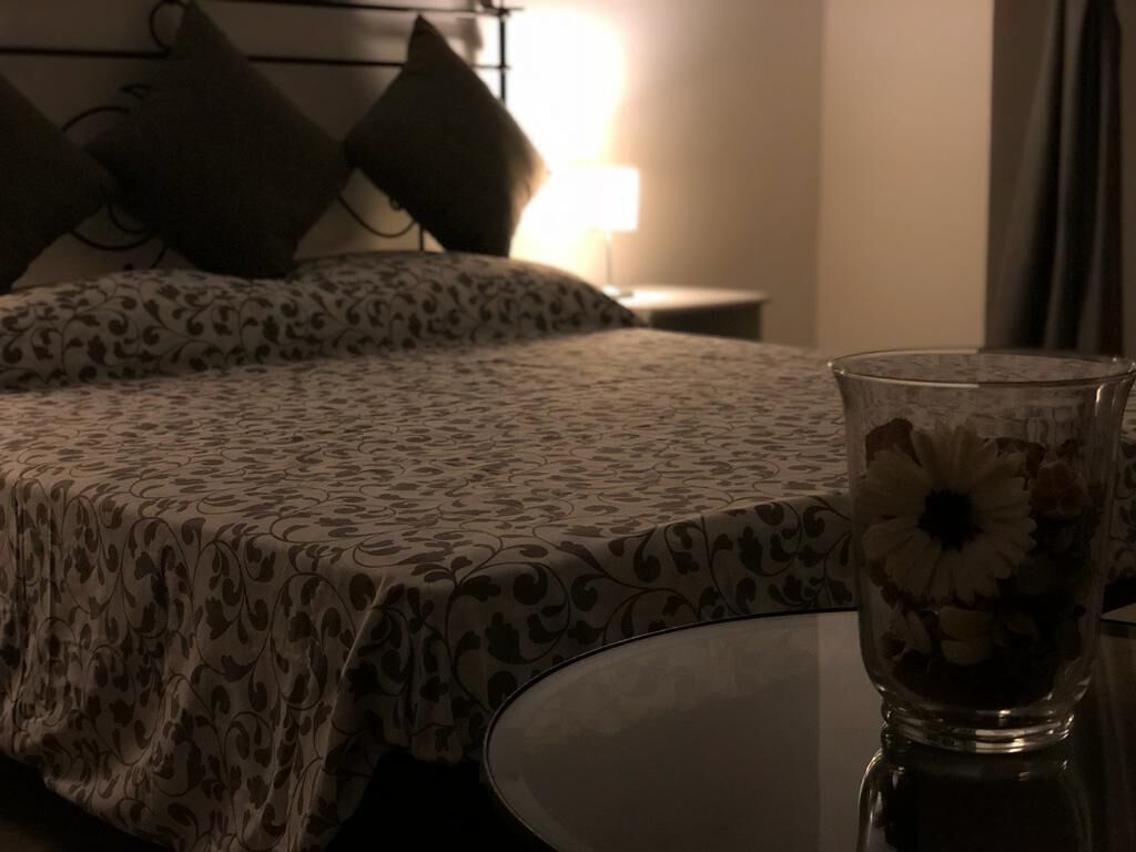 A bed with a patterned bedspread, a bedside table with a glass vase of flowers, and a lamp in the background.