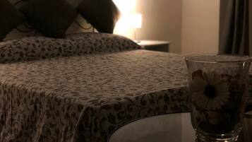 A bed with a patterned bedspread, a bedside table with a glass vase of flowers, and a lamp in the background.