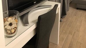 Un salon moderne avec un téléviseur à écran plat posé sur un meuble blanc, un vase en verre contenant des fleurs séchées, un canapé gris et un rideau gris.