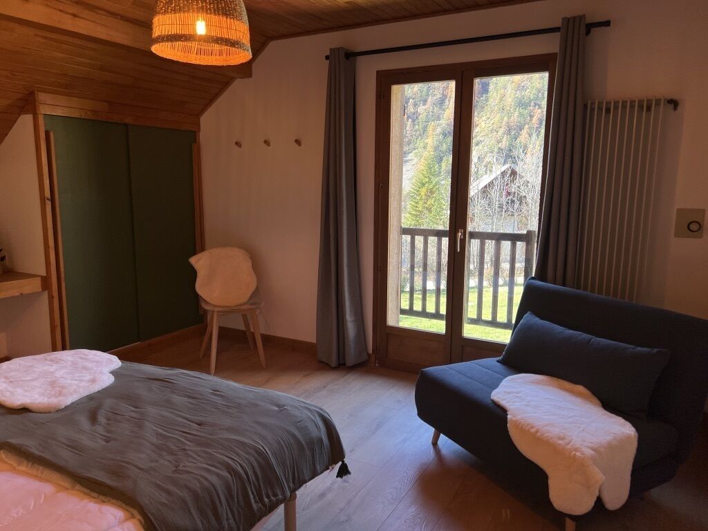 Une pièce comprenant un lit, un canapé sombre, un fauteuil et donnant sur un balcon avec une balustrade en bois.