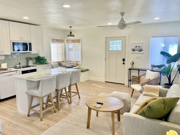 A modern kitchen with white cabinets, a microwave, and a dining area with stools. There is a living room with a sofa and a coffee table.