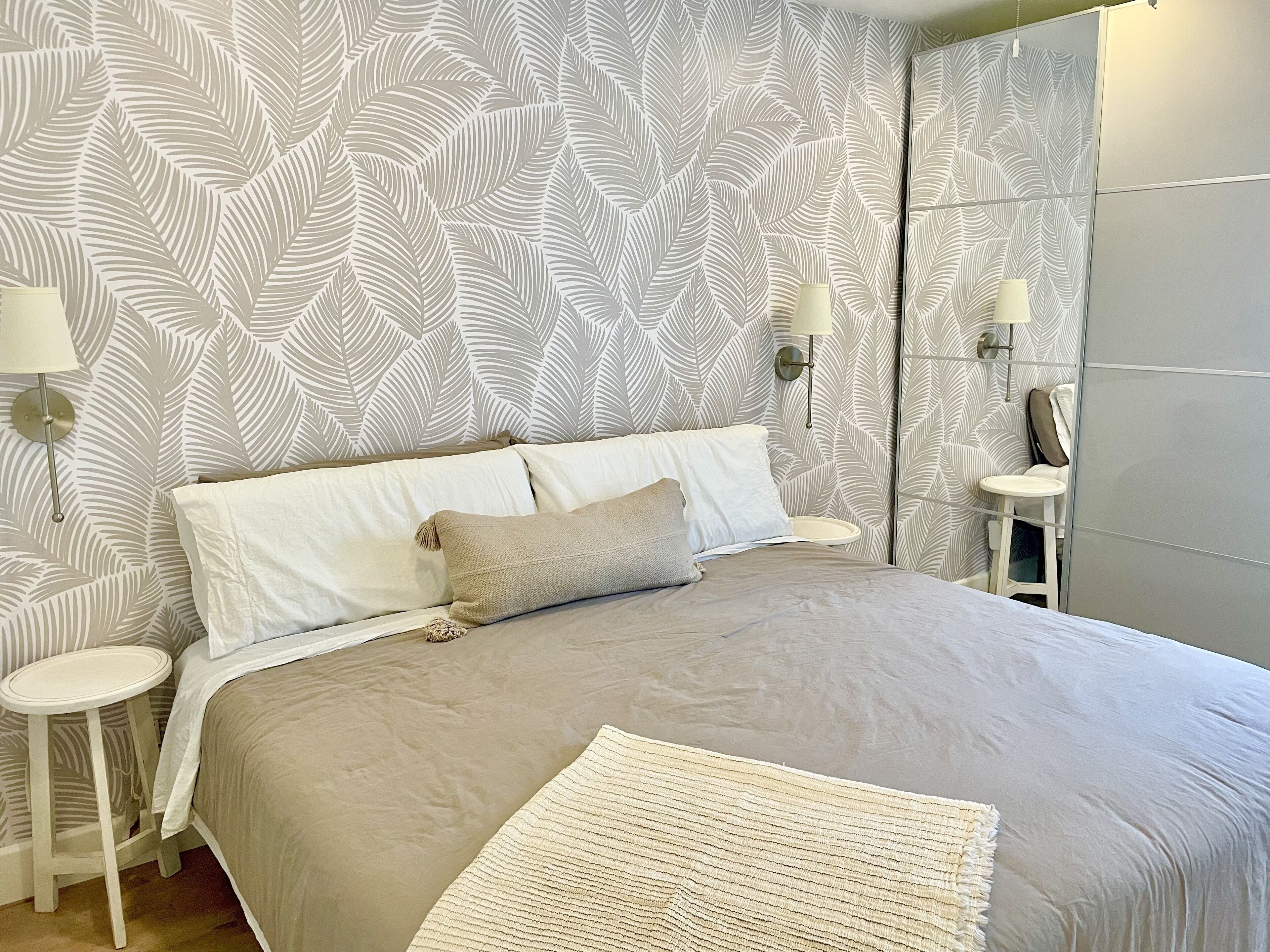 A neatly made bed with a beige comforter and pillows, a white throw blanket, and a patterned wallpaper with leaf designs. There are two bedside tables with lamps on either side of the bed.