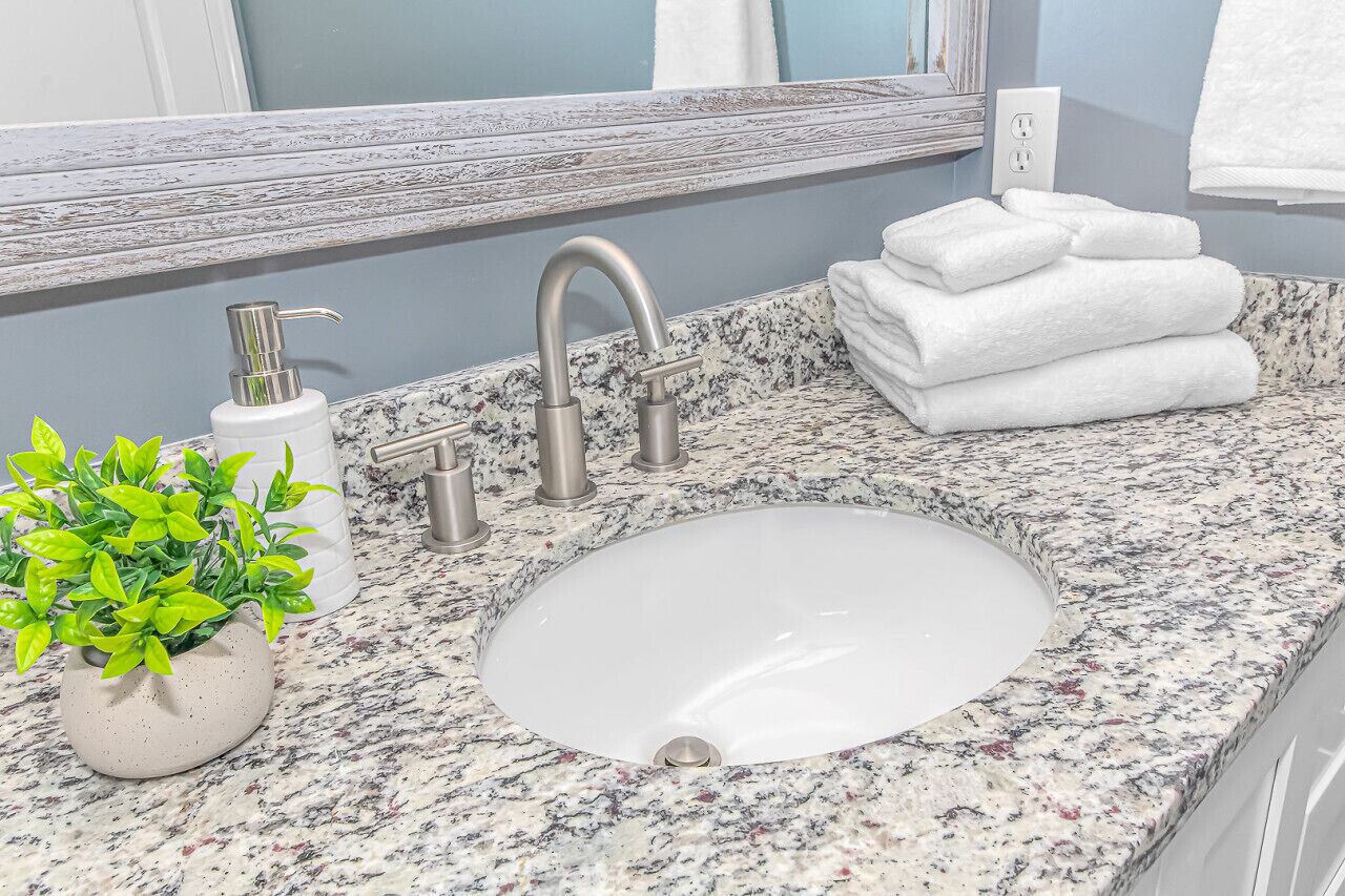 A bathroom with a granite countertop, a white sink, a silver faucet, a potted plant, and a stack of white towels.