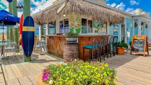 A beachside restaurant with a thatched roof, outdoor seating, and a sign advertising local craft beers. - Grand Strand 5BR Retreat | Heated Pool | Steps to Garden City Beach (Murrells Inlet)