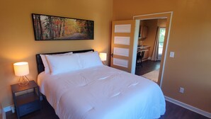 A bedroom with a large bed, a nightstand with a lamp, a ceiling light fixture, and a door leading to another room. - Deluxe Queen with Park View (Laurium)