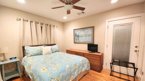 A bedroom with a bed, a nightstand, a dresser, a television, a ceiling fan, and a door.