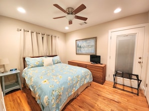 A bedroom with a bed, a nightstand, a dresser, a television, a ceiling fan, and a door.