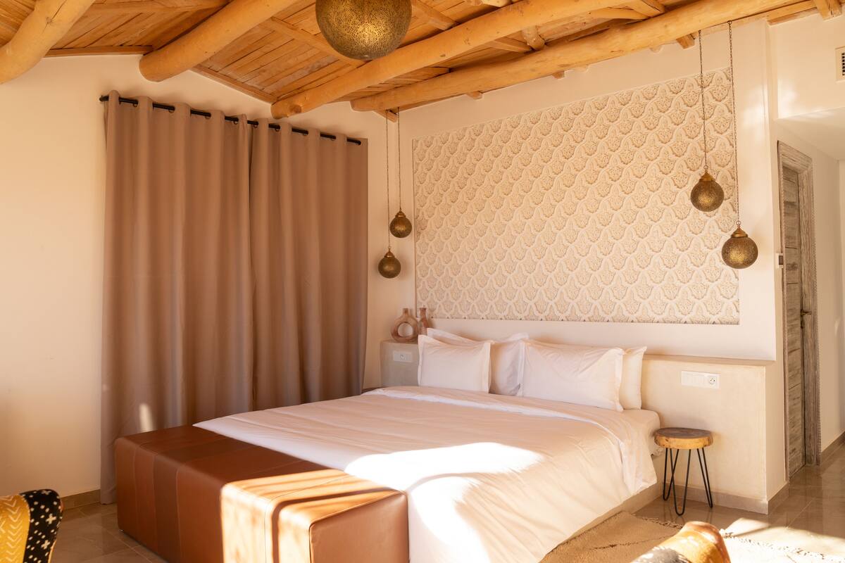 A bedroom with a wooden ceiling, a patterned wall, a bed with white bedding, a small bedside table, and a brown ottoman.