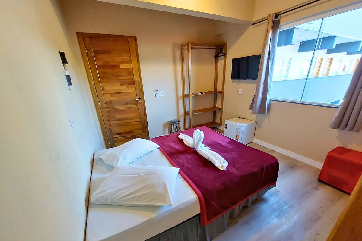 Une petite chambre à coucher, bien rangée, avec un lit, une armoire en bois, un petit tabouret, une télévision et une fenêtre avec des rideaux.