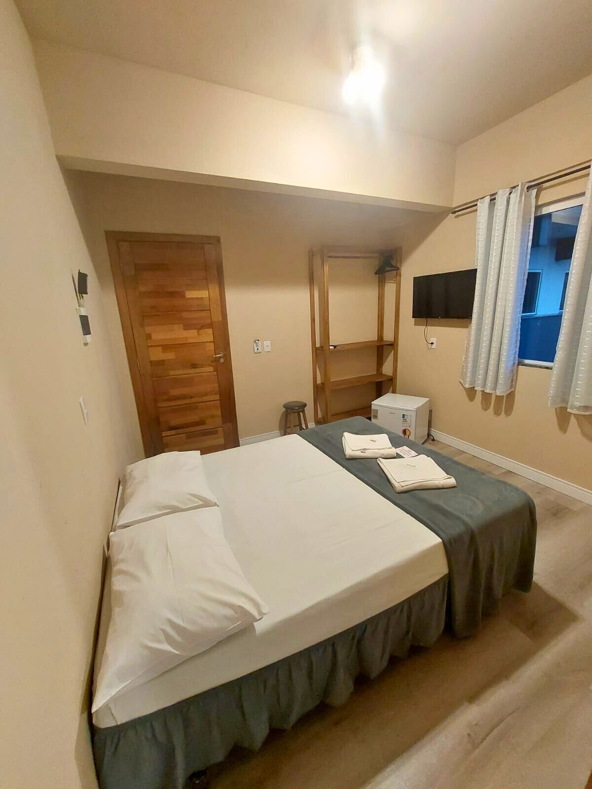 Un dormitorio con cama, una puerta de madera, televisor y ventana con cortinas.