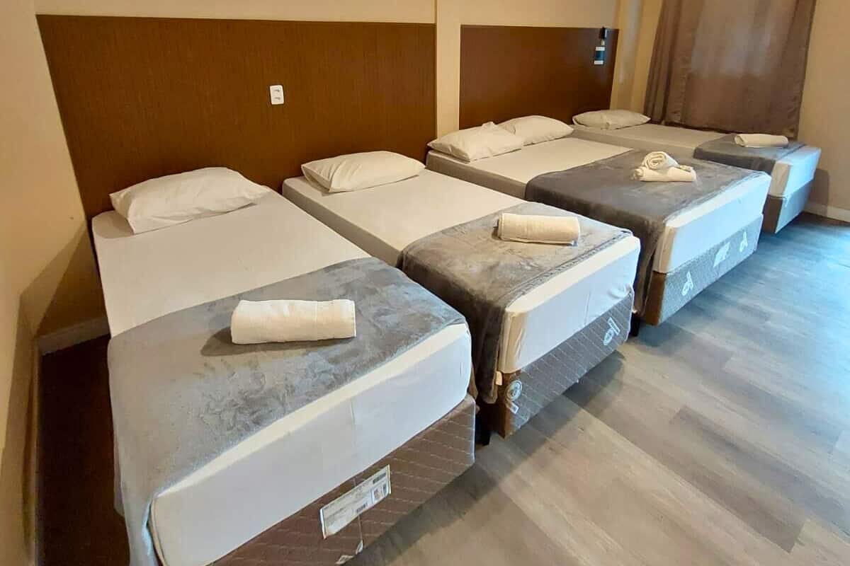 Une chambre d’hôtel avec quatre lits simples, chacun équipé d’un oreiller et d’une serviette enroulée. La chambre est dotée d’un plancher en bois et d’une tête de lit marron.