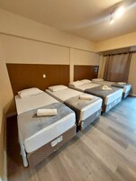 Quarto de hotel com quatro camas de solteiro, cada uma com almofada e toalha enrolada. O quarto possui piso de madeira e cabeceira marrom.