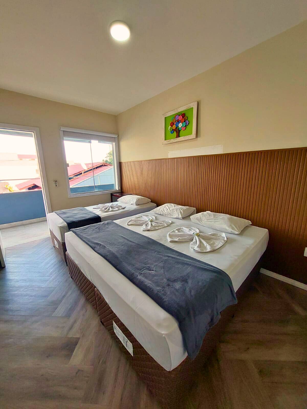 A hotel room with two beds, a wooden headboard, a painting of a tree, and a view through the window.
