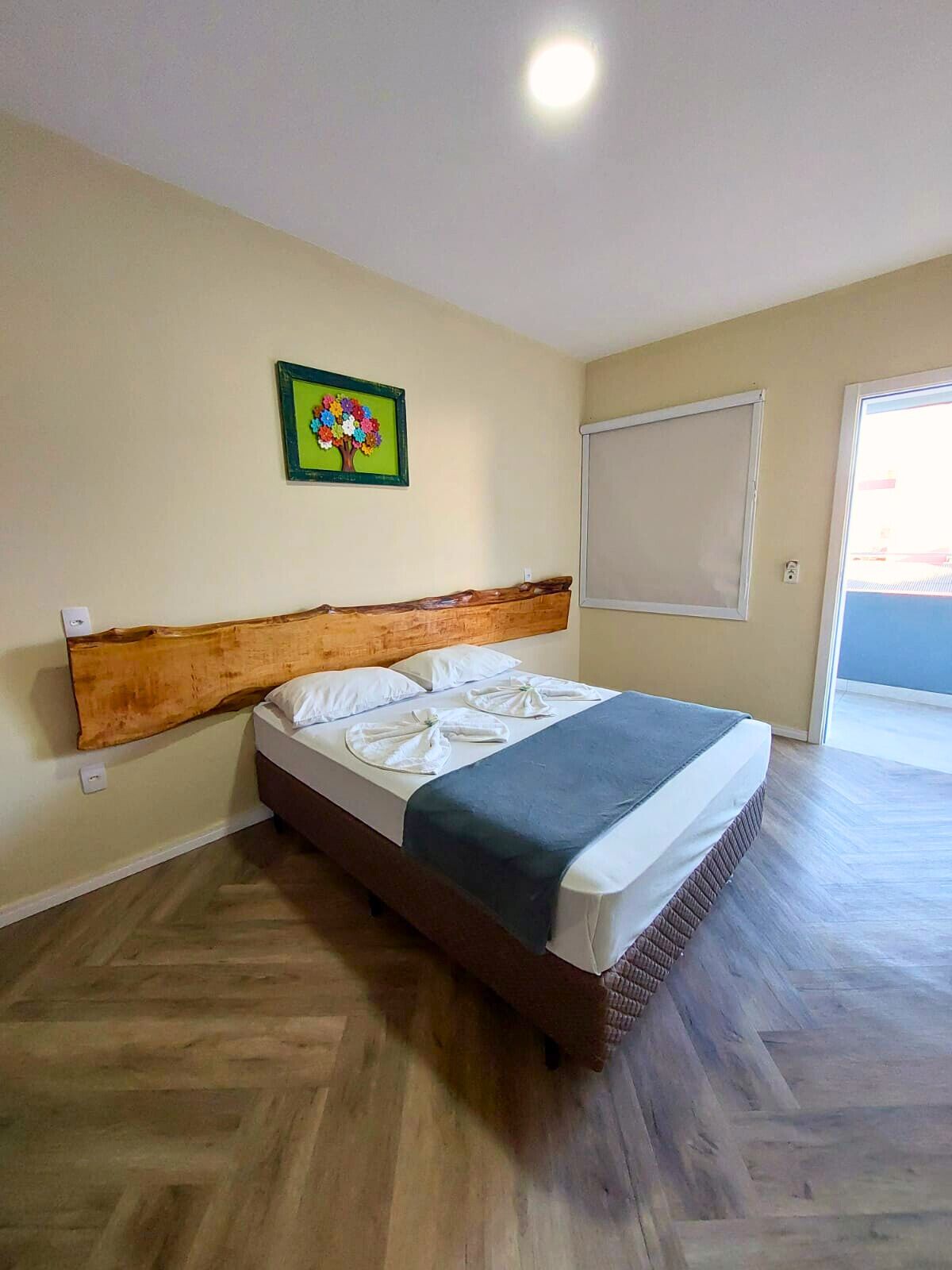 A bedroom with a wooden headboard, a bed with a blue blanket, a green-framed picture on the wall, and a window with a white blind.
