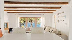 Une pièce avec une table blanche, un canapé avec des coussins, des poutres en bois au plafond et une fenêtre donnant sur des arbres et l’eau.