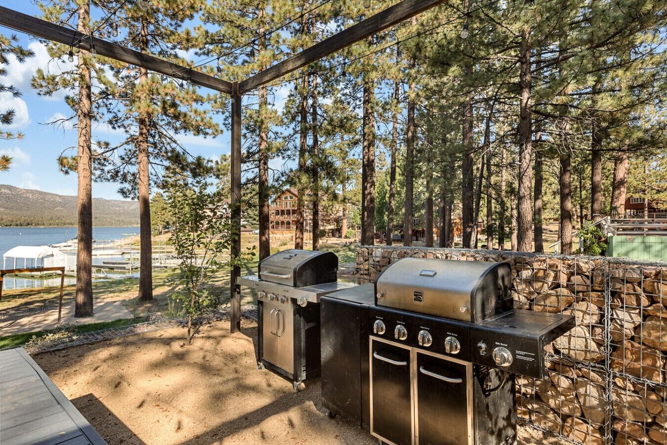 An outdoor grill area with a stone wall and wooden pergola, surrounded by trees and a lake view.