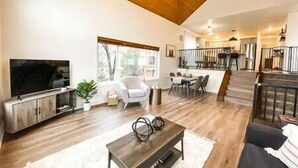 Ein großzügiges Wohnzimmer mit Flachbildfernseher, einem weißen Sofa und einem Holztisch. Es gibt eine Essecke mit Tisch und Stühlen, und eine Treppe, die zu einer oberen Ebene führt.