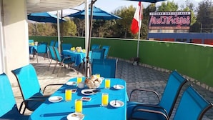 An outdoor patio with blue tables and chairs, some with breakfast items and orange juice. A sign for 'MULTIFUEGOS' is visible in the background.