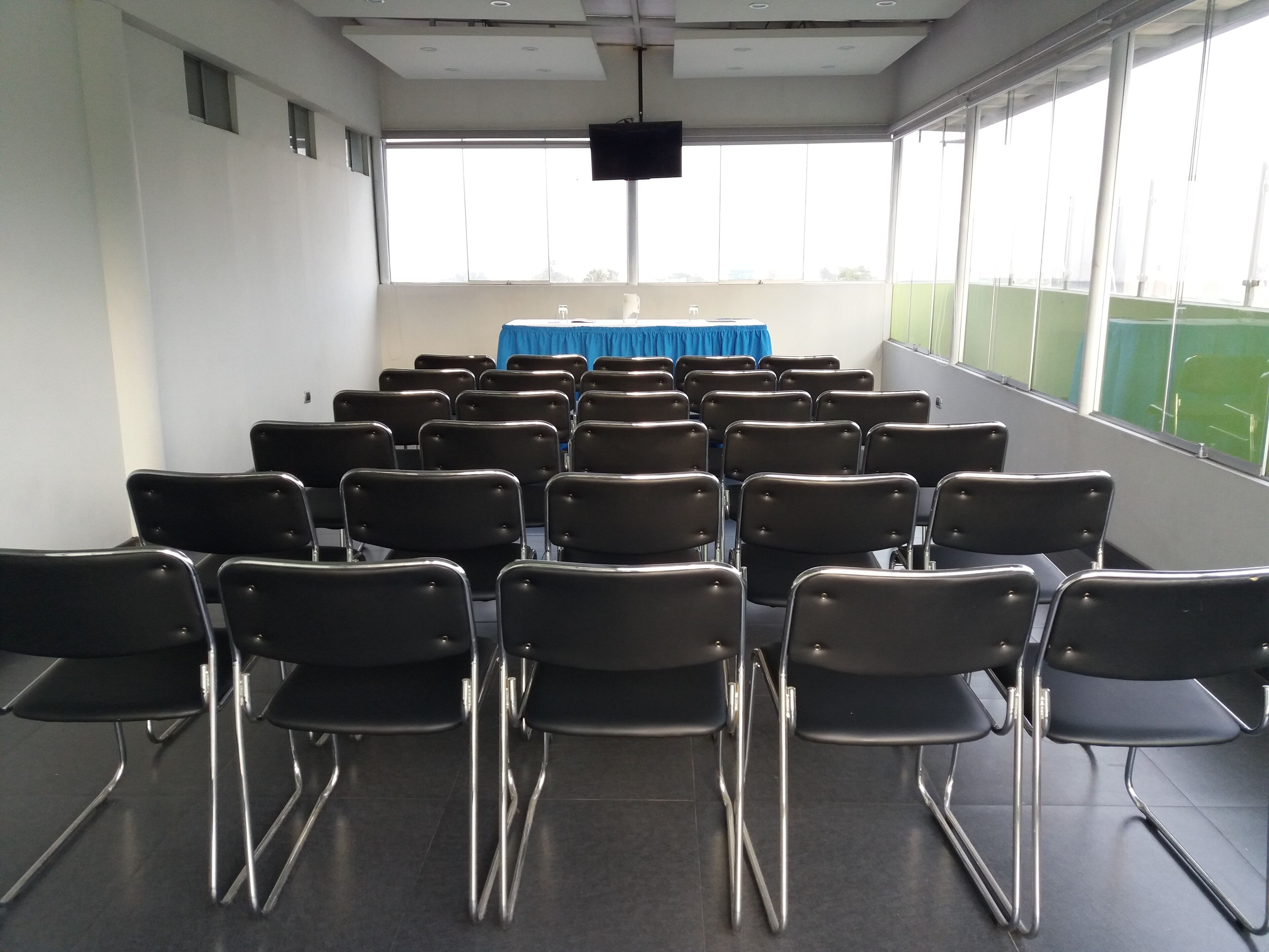 Uma sala de conferências com fileiras de cadeiras, uma mesa azul na frente e grandes janelas.