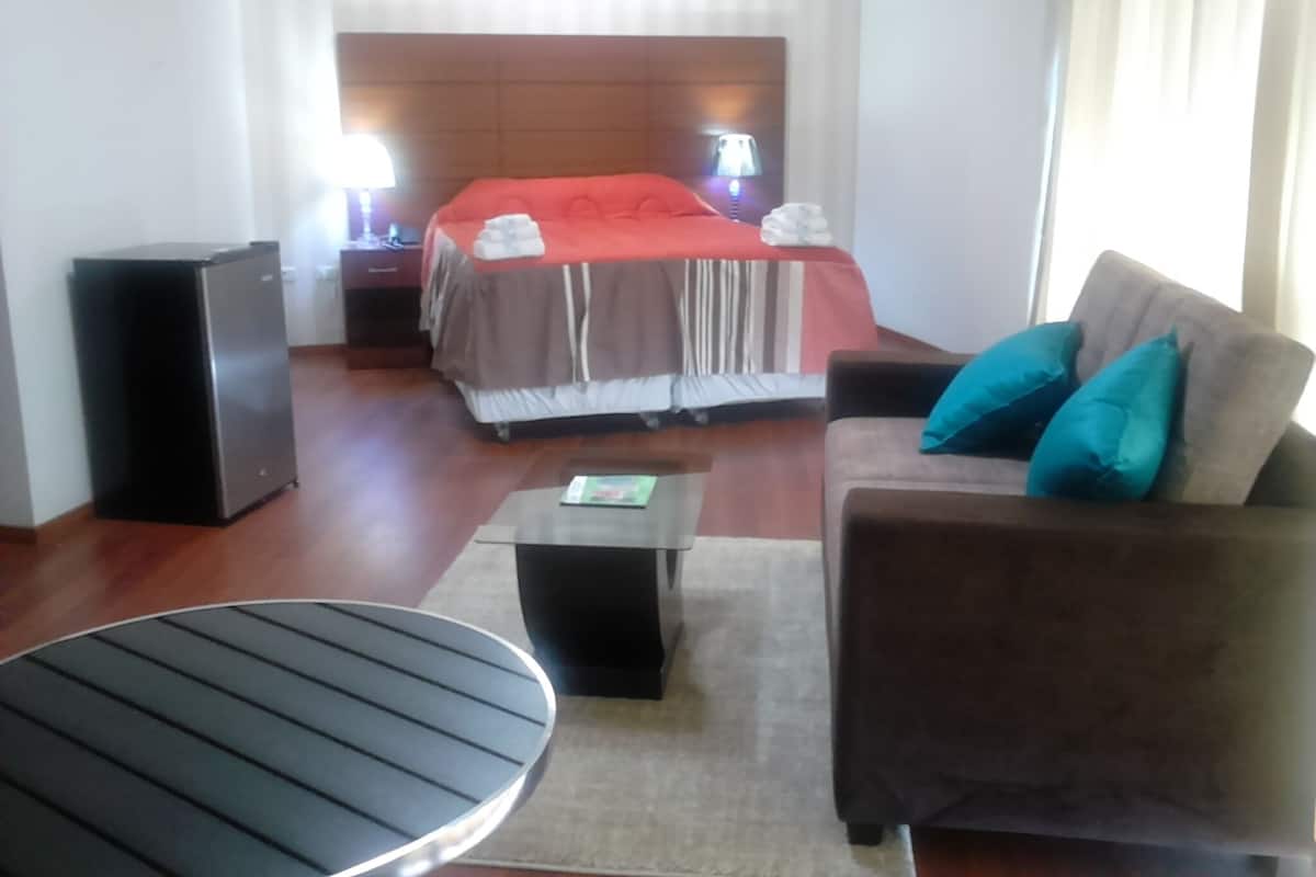 A hotel room with a bed, a sofa, a coffee table, a TV, and a speaker.