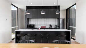 Una cocina moderna con una isla grande, taburetes negros tipo bar, iluminación colgante y un refrigerador de acero inoxidable.