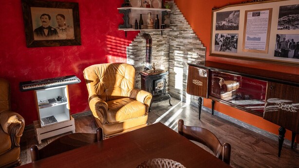 A room with a red wall, a vintage chair, a wooden cabinet, a stone fireplace, and a framed photograph.