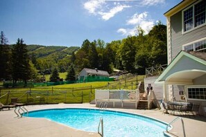 En swimmingpool med terrasse, en overdækket patio og udsigt til et grønt hus og skovklædte bakker i baggrunden.