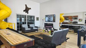 A living room with a shuffleboard table, sofas, a TV, and a kitchen area with yellow spiral slides. - Experience Scottsbluff charm in our spacious 4-bedroom retreat (Scottsbluff)