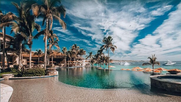 A resort pool with palm trees, a clear blue sky, and boats in the background. - Hacienda Beach Club, Beachfront, 5-Star, Luxury (Cabo San Lucas)
